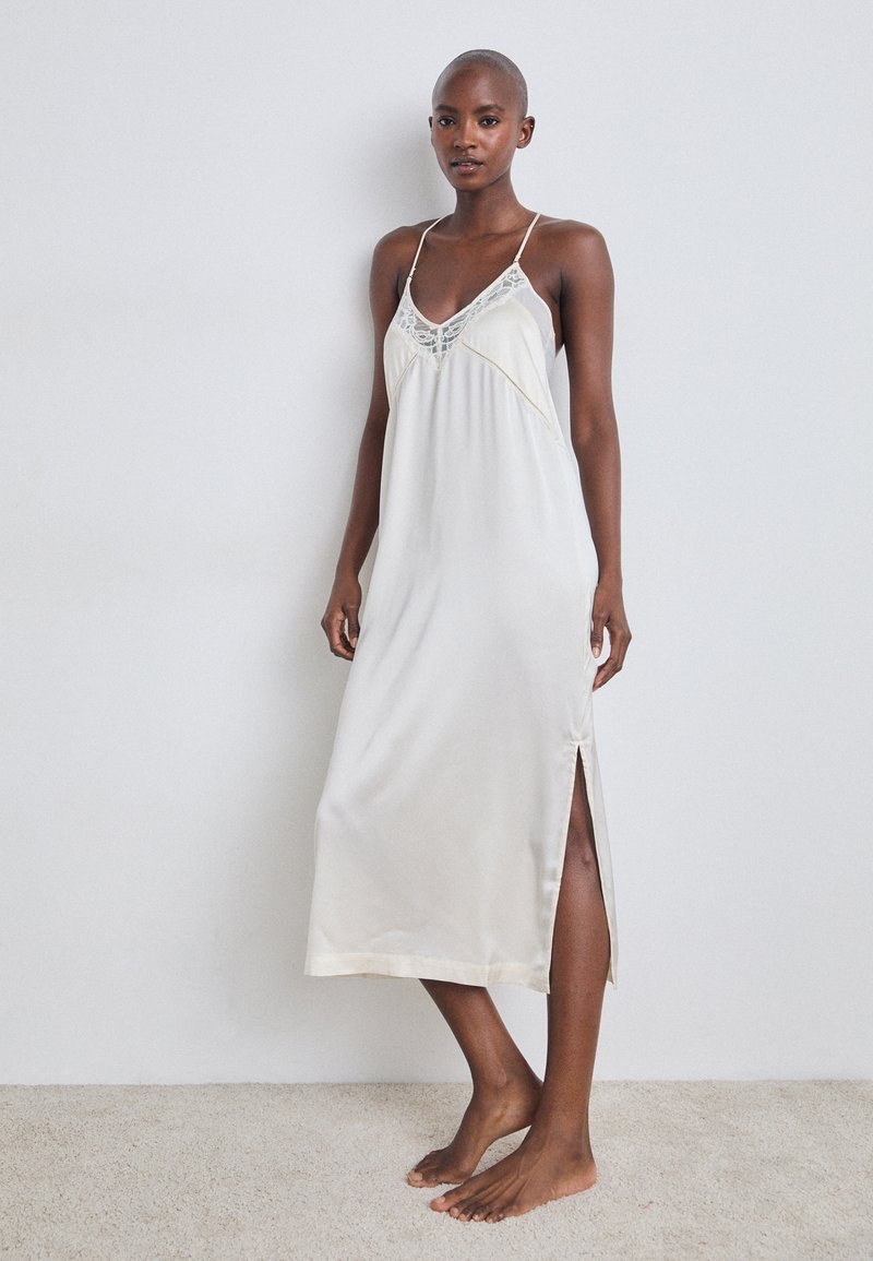 Κρέμα σατέν φόρεμα με κοπή slip, με κεντημένο τελείωμα στο λαιμό, ρυθμιζόμενες τιράντες spaghetti και λεπτομέρεια με σχισμή στο πλάι. Λείο, ρέον ύφασμα.