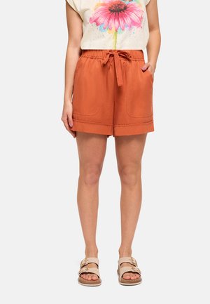 Mujer vistiendo pantalones cortos de color naranja óxido con cordón y bolsillos delanteros, camiseta blanca con un gran estampado de flor rosa y sandalias beige con hebilla.