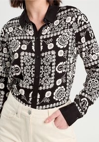 Chemise boutonnée noire et blanche avec motifs floraux, dotée d'un col classique, de manches longues et de poignets noirs. Matière en coton.