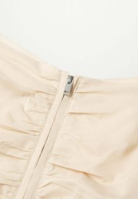 Vestido beige con cierre de cremallera y herrajes plateados, y textura de tejido fruncido cerca del busto, que resalta una superficie suave.