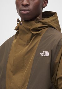Άνδρας που φοράει δίχρωμο καφέ μπουφάν North Face με κουκούλα και λογότυπο στο στήθος, κοιτάζοντας ελαφρώς προς τα δεξιά με φόντο απλό.