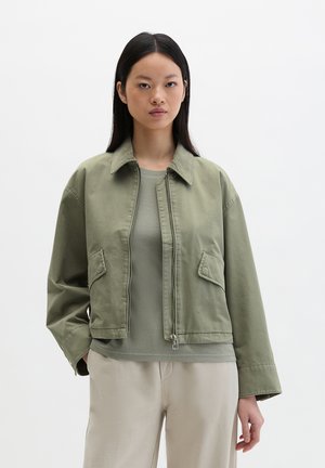 Jeune femme portant une veste courte vert olive par-dessus un haut vert assorti et un pantalon beige, debout devant un fond blanc uni.