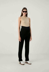Haut sans manches en maille beige, pantalon noir taille haute, baskets blanches avec accents noirs et lunettes de soleil noires. Textures lisses partout.