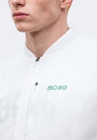 Κοντινό πλάνο του κάτω μέρους του προσώπου και του λαιμού ενός ατόμου που φοράει λευκή μπλούζα με φερμουάρ και πράσινο κείμενο "BORG" στο στήθος.