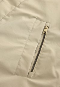 Beige pufferjakke med en lynlåse-lomme. Stoffet er glat med synlige stikninger; lynlåsdetaljerne er i en mørkere nuance.