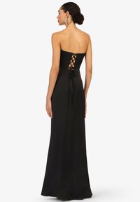 Femme ayant les cheveux bruns dans un chignon bas, vêtue d'une robe de soirée noire sans manches avec un laçage dans le dos, se tenant devant un fond blanc.