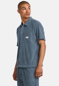 Camisa de manga corta, con textura en azul apagado, que presenta un patrón floral, cierre de botones, un bolsillo en el pecho y una marca sutil.