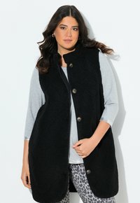 Nainen, jolla on pitkät tummat hiukset, käyttää hopeisia rengaskorviksia, mustaa nappivillaista liivimekkoa vaaleanharmaan paidan päällä, ja harmaita leopardikuvioisia housuja.