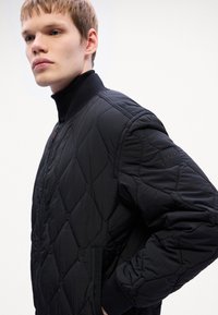 Προβληματισμένο μαύρο μπουφάν bomber με φερμουάρ, ραμμένο γιακά και λογότυπο στη μανσέτα. Διαθέτει υφή καθώς και πλευρικές τσέπες.