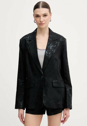 Donna con capelli tirati indietro, che indossa orecchini a cerchio argento, blazer nero testurizzato e pantaloncini abbinati, su uno sfondo chiaro e uniforme.