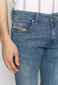 Blue denim jeans with a slim fit, featuring a visible pocket with a label, subtle fading, and yellow stitching accents.