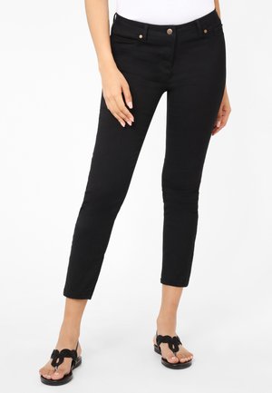 Black skinny jeans with a mid-rise waist, featuring traditional pockets and metal button closures, paired with black sandals with a bow accent.