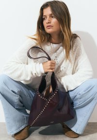 Deep burgundy leather bag with studded accents, slouchy design, and a single long strap, held by a person squatting, wearing casual attire.