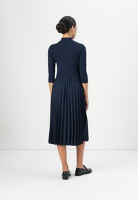 Navy pleated dress with a fitted ribbed top and three-quarter sleeves, featuring a knee-length skirt and black slip-on shoes.