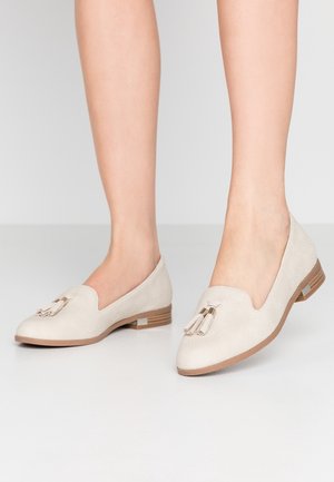 Pair of beige textured loafers with tassels worn on feet, featuring low wooden heels and rounded toes on a smooth light surface.