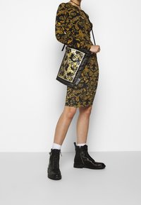 Svart och guldmönstrad handväska i rektangulär form, med ett platt handtag och strukturerad design. Kombinerad med en åtsittande svart och guldklänning samt svarta stövlar.