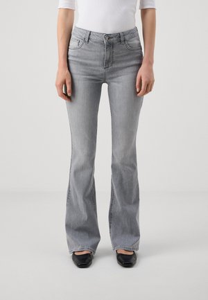 Kobieta w jasnoszarych rozszerzanych dżinsach, czarnych butach o kwadratowym nosku i białym dopasowanym topie, stojąca na tle jednolitego jasnego tła.