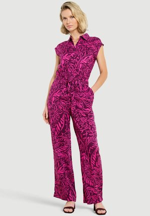 Vrouw die een paarse jumpsuit draagt met felroze bladpatronen, korte mouwen, een getailleerde taille en zwarte open-toe sandalen.