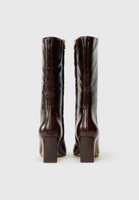 Un par de botas altas de cuero marrón oscuro con tacones en bloque y cremalleras doradas en la parte trasera, mostradas desde la parte trasera sobre un fondo blanco.