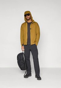 Világosbarna, kapucnis cipzáras dzseki, fekete póló, fekete nadrág és fekete hátizsák. A dzsekin kis logó és oldalzsebek találhatók.