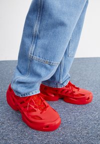 Červené tenisky s lehkým a prodyšným designem, zdobené síťovinovými detaily a tlustými podrážkami, v kombinaci se světle modrými džínami.