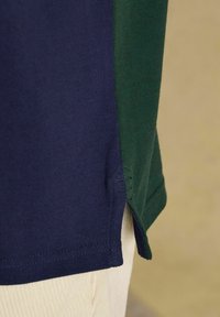 Nahaufnahme des Saums eines marineblauen und grünen Poloshirts mit seitlichem Schlitz, getragen über beigefarbenen Cordhosen.