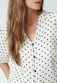 Weißes Bluse mit schwarzen Punkten, Knopfverschluss vorne, breiter Kragen und kurzen Ärmeln. Texturierter Stoff mit glatter Oberfläche.