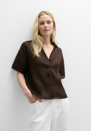 Mujer rubia con camisa de manga corta marrón oscuro abotonada y pantalones blancos, de pie con las manos en los bolsillos frente a un fondo blanco.