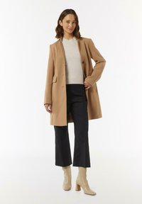 Camel-wollmischung Mantel mit einem einzelnen Knopfverschluss, mit Reverskragen und zwei seitlichen Taschen, getragen über einem hellgrauen Oberteil und navyblauen Hosen.