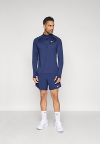 Μπλε ναυτικό αθλητικό μπλουζάκι με μακριά μανίκια και φερμουάρ στον γιακά, συνδυασμένο με μπλε ναυτικά σορτς. Και τα δύο έχουν λευκό λογότυπο Nike. Το σύνολο είναι κατασκευασμένο από ελαφρύ, αεριζόμενο υλικό.