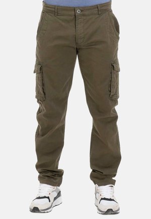 Homme portant un pantalon cargo vert olive et des baskets blanches, debout devant un fond uni, jambes légèrement écartées.