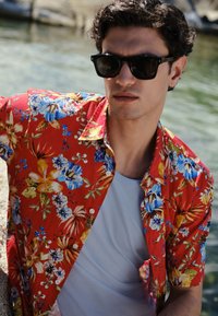 Jonge man met een zwarte zonnebril en een rood bloemenoverhemd over een wit T-shirt, zittend bij een waterpartij.