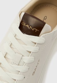 Fehér bőrből készült sportcipő fehér fűzőkkel és barna bőr nyelvvel, amelyen arany betűkkel látható a "GANT" felirat a nyelven és az oldalán.