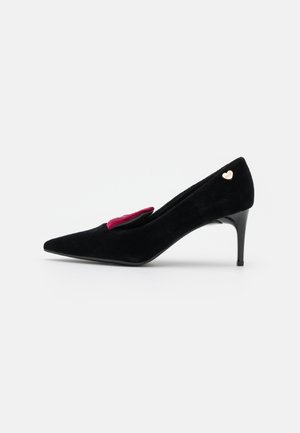 Chaussure à talon haut en daim noir à bout pointu, talon fin, petit détail cœur doré à l’arrière et nœud rouge sur le dessus à l’avant.