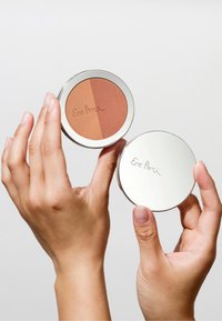 Rund blush-kompakt som viser to nyanser: en ferskenfarget og en dypere brun. Sølvmetallhylse med merkevarenavnet inngravert på lokket.