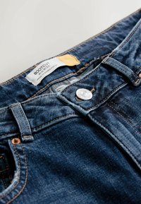 Blugi bootcut din denim albastru închis cu talie medie. Caracteristicile includ un nasture metalic, cusături în contrast și o etichetă inscripționată "BOOTCUT".