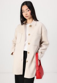 Beige Wollmischung Hemdjacke mit zwei Vorderaschen und goldfarbenen Knöpfen, kombiniert mit einer strukturierten roten Lederhandtasche.