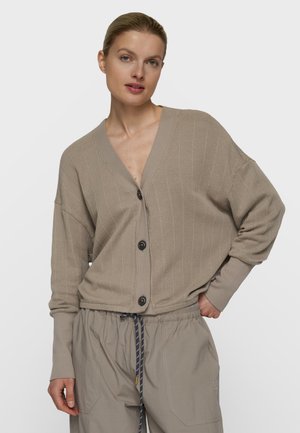 Kvinde iført en løs beige knappet cardigan og matchende lyse beige snøre bukser, poserer med den ene hånd i lommen mod en ensfarvet baggrund.
