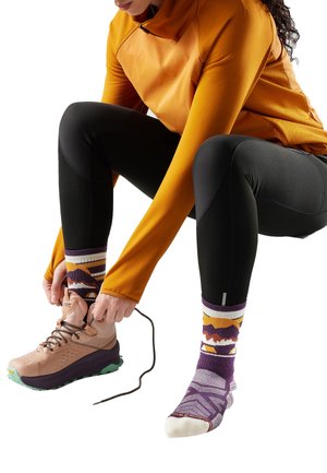 Person iført orange langærmet top og sorte leggings, der binder snørebåndene på en vandrestøvle, med farverige mønstrede sokker synlige.