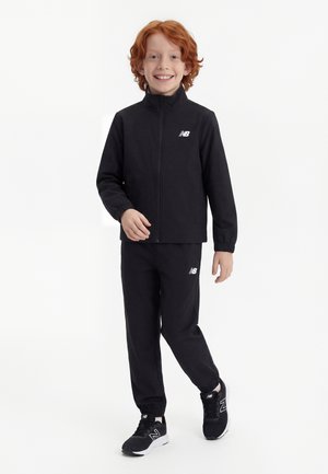 Uśmiechnięte dziecko z rudymi włosami, ubrane w czarny dres New Balance i czarne trampki, stojące na białym tle.