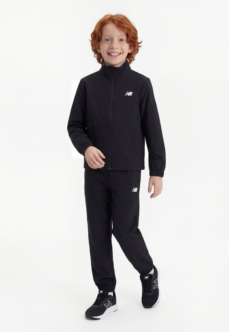 Χαμογελαστό παιδί με κόκκινα μαλλιά, φορώντας μαύρη φόρμα New Balance και μαύρα αθλητικά παπούτσια, στέκεται μπροστά σε λευκό φόντο.