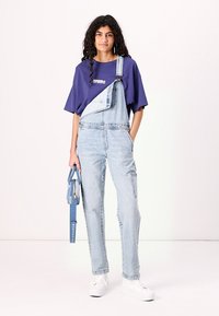 Salopette en denim bleu clair avec une seule bretelle, associée à un T-shirt violet ample et des baskets blanches. Comprend un sac bleu.