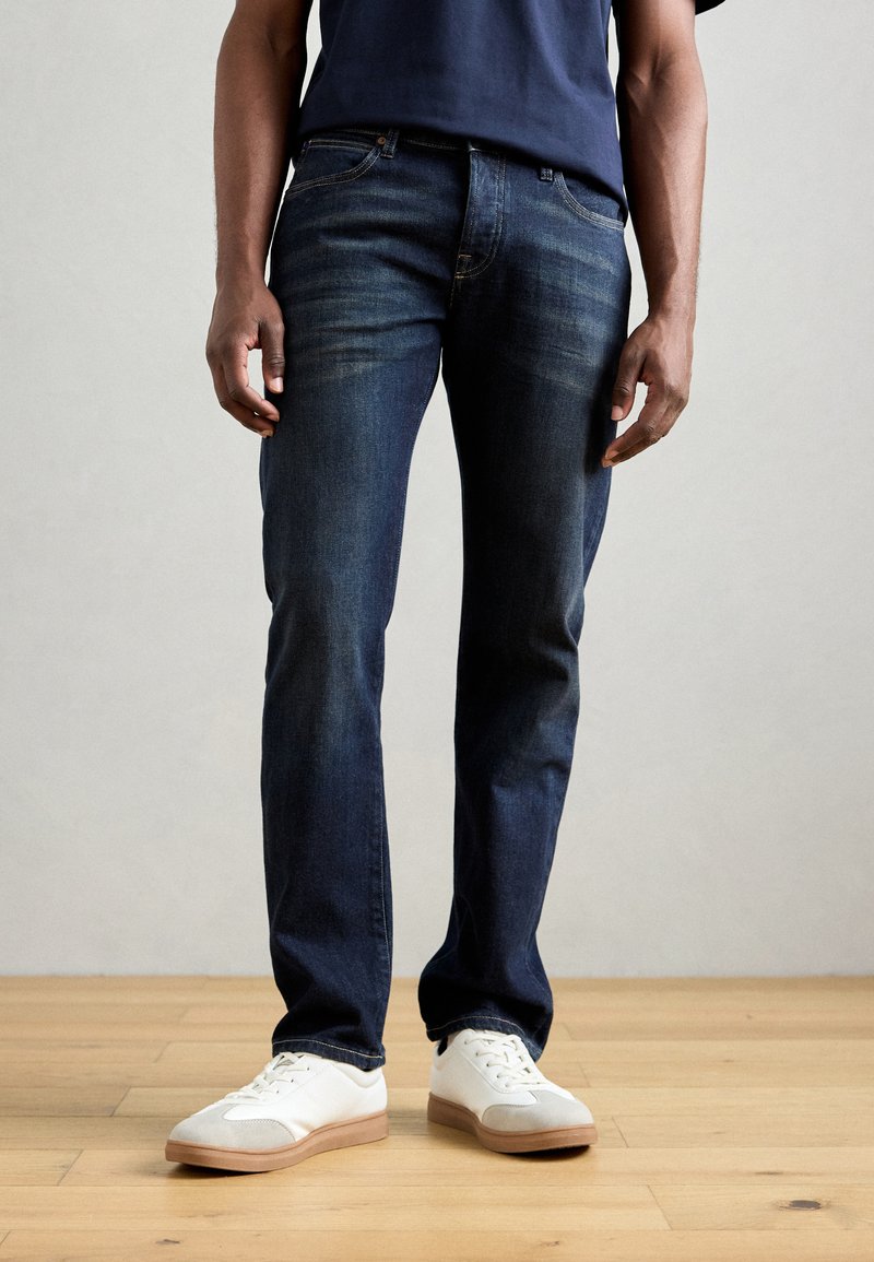 Dunkelblaue Jeans mit geradem Schnitt, dezentem Verblassen und klassischem Fünf-Taschen-Design, kombiniert mit weißen Sneakers auf einem Holzboden.