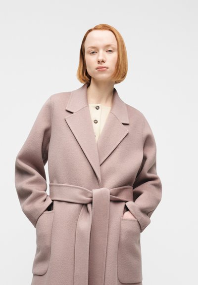 Νεαρή γυναίκα με κοντά κόκκινα μαλλιά, φοράει ροζ παστέλ μάλλινο παλτό με ζώνη, πέτο και μεγάλες μπροστινές τσέπες, με τα χέρια μέσα στις τσέπες.