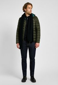 Grön pufferjacka med dragkedja fram, svart scarf, svart pikétröja, mörka jeans och svarta skor. Modellen står mot en neutral bakgrund.