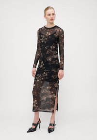 Robe longue noire en tissu transparent, ornée de motifs floraux, avec des manches longues et des fentes sur les côtés. Portée avec des escarpins noirs à lanières.