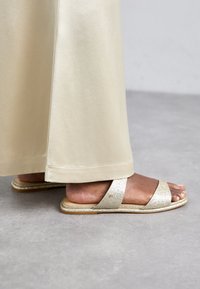 Glittery gold slide sandals with two overlapping straps and a flat sole. Worn with a light beige satin skirt.
