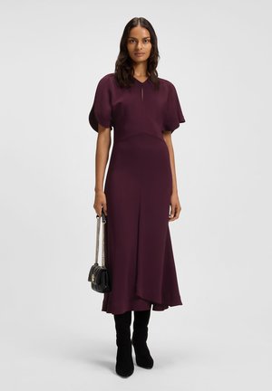 Woman wearing a sleeved plum midi dress, black knee-high boots, and holding a small black handbag with gold chains.