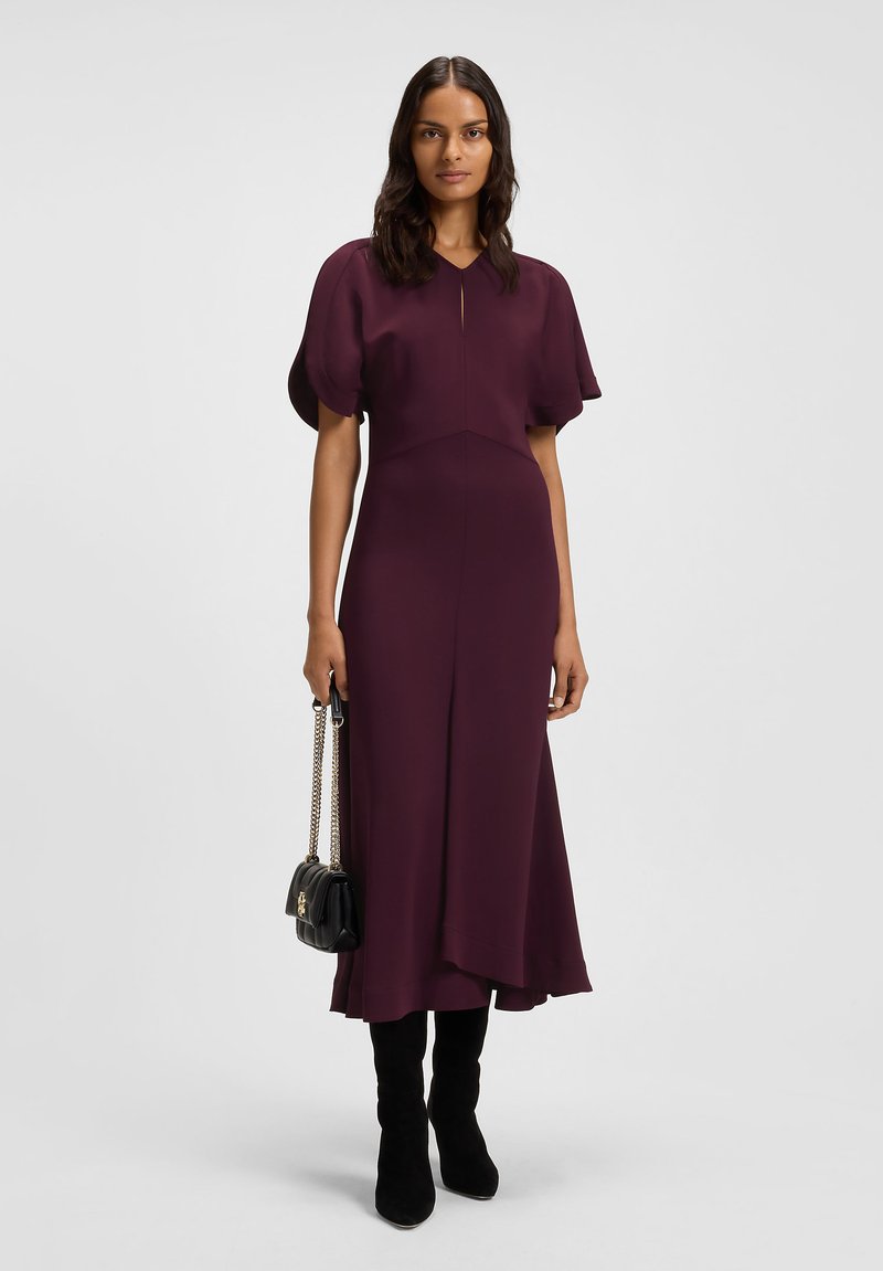Donna che indossa un vestito midi prugna con maniche, stivali neri fino al ginocchio e tiene in mano una borsa nera con catene dorate.