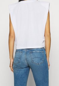 Biały krótki top na ramiączkach z szerokimi wycięciami po bokach, w połączeniu z jasnoniebieskimi dżinsami z tylnymi kieszeniami i delikatnym przetarciem.